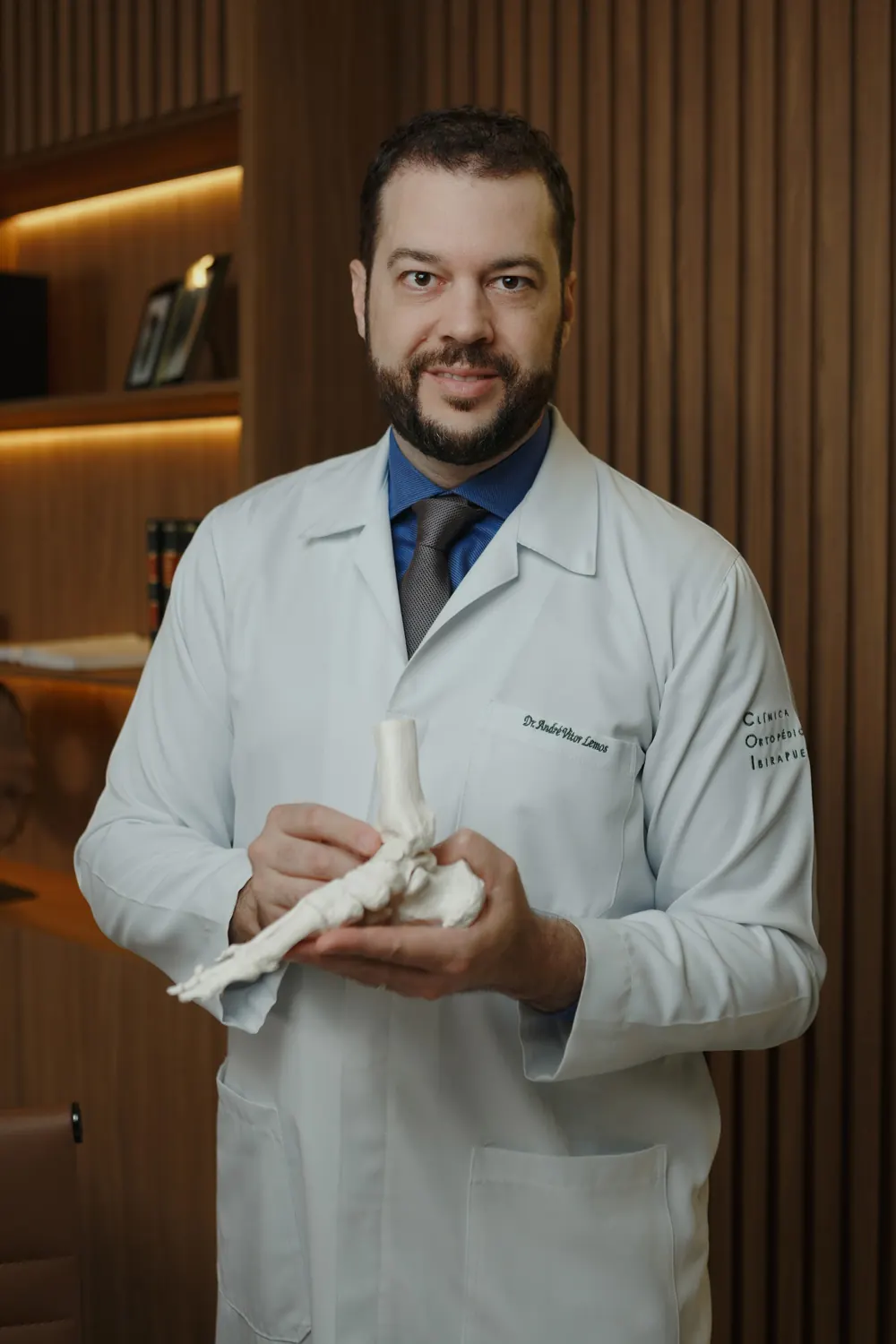 Dr. André Lemos no consultório, segurando um modelo anatômico do pé e do tornozelo